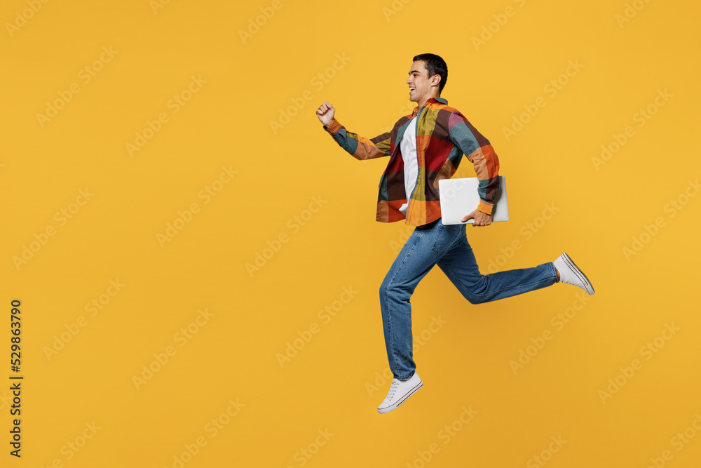 Full body young middle eastern IT man 20s in casual shirt white t-shirt jump high hold closed laptop pc computer run fast isolated on plain yellow background studio portrait People lifestyle concept