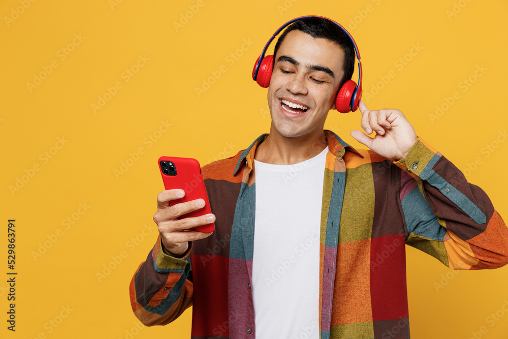 Young happy middle eastern man 20s wear casual shirt white t-shirt headphones listen to music hold use mobile cell phone isolated on plain yellow background studio portrait People lifestyle concept Young happy middle eastern man 20s wear casual shirt white t-shirt headphones listen to music hold use mobile cell phone isolated on plain yellow background studio portrait People lifestyle concept