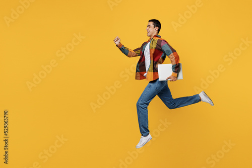 Full body young middle eastern IT man 20s in casual shirt white t-shirt jump high hold closed laptop pc computer run fast isolated on plain yellow background studio portrait People lifestyle concept