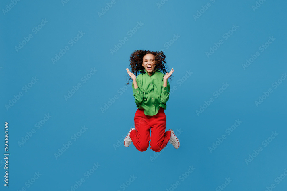 Full body excited fun young woman of African American ethnicity 20s she wear green shirt jump high spread hands look camera isolated on plain blue background studio portrait. People lifestyle concept. Full body excited fun young woman of African American ethnicity 20s she wear green shirt jump high spread hands look camera isolated on plain blue background studio portrait. People lifestyle concept.