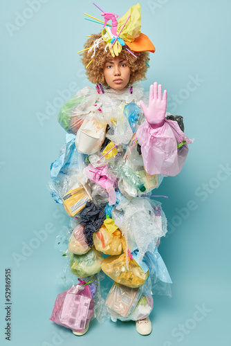 Serious confident woman keeps palm raised towards camera makes forbidden gesture demands to stop polluting environment realizes seriousness of big environmental problem stands against blue wall