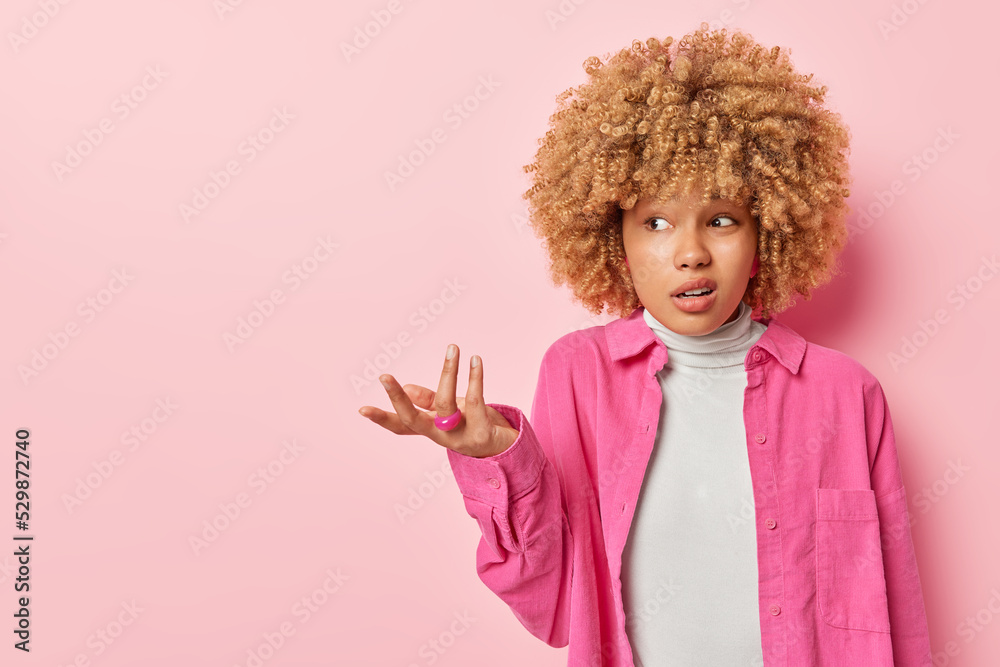 Horizontal shot of confused woman shrugs shoulders has questioned puzzled expression purses lips dressed in shirt isolated over pink background empty space for your advertising content. So what