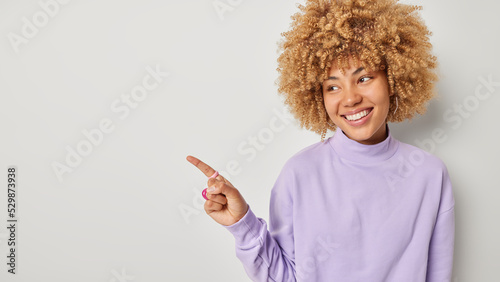 Horizontal shot of happy beautiful woman with curly blonde hair points index finger aside on blank space shows advertisement smiles pleasantly isolated over grey background. Look at this promo