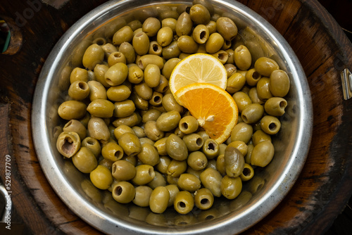 Green olives on display in a silver bowl on a stall in a market place where fresh food is bought by people who like healthy Mediterranean food. The tasty nibble is garnished with range and lemon