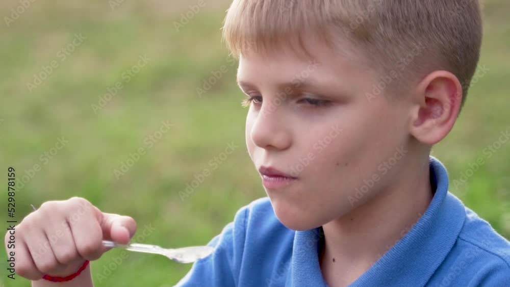 A blond-haired boy in a blue T-shirt eats ice cream with a plastic spoon in nature. Close-up. Slow motion. A blond-haired boy in a blue T-shirt eats ice cream with a plastic spoon in nature. Close-up. Slow motion.