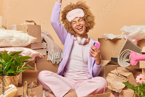 Overjoyed woman with curly hair wears casual clothes and sleepmask holds smartphones listens music has upbeat mood surrounded by cardboars boxes full of personal belongings relocates to apartment photo