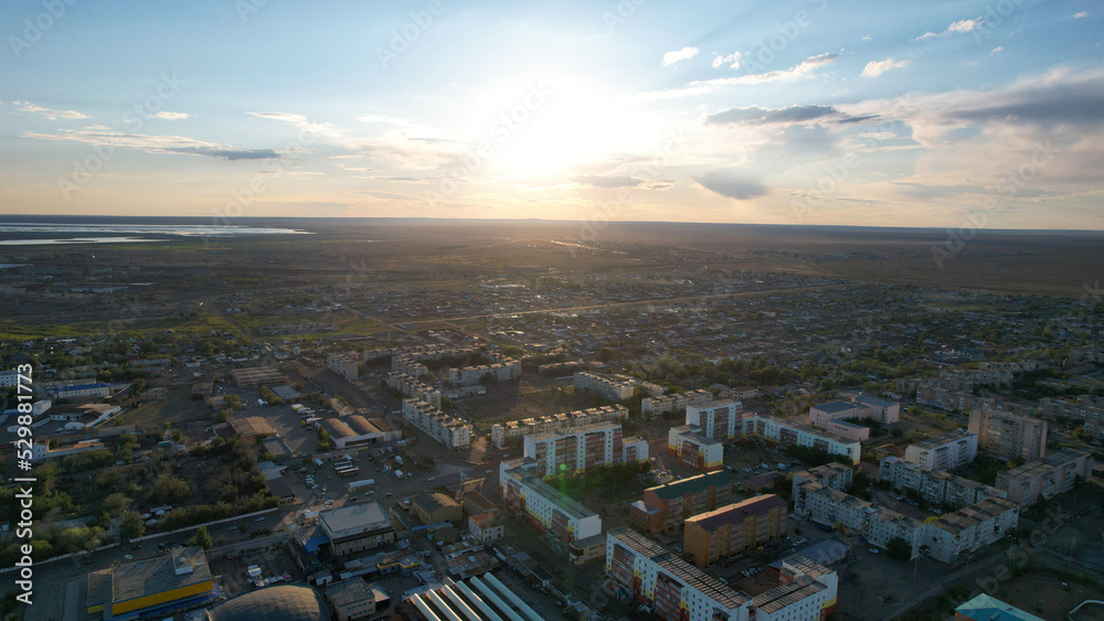 Bright sun with clouds before sunset over the city. Dark white clouds in a blue sky. A small town and Lake Balkhash. Green trees, roads with moving cars and high-rise buildings. Around the steppe