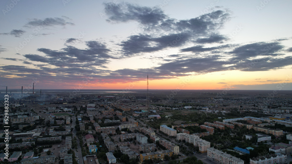 Orange sunset over a small town. Top view from a drone. Blue-purple clouds against background of houses, a smoking factory and a TV tower. Cars drive. There is a bright light at the metal drain plant