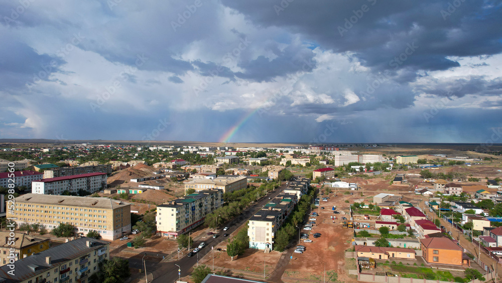 Bright rainbow and rain over the city of Balkhash. The small town is all wet. Large clouds of white-blue color float by. Cars drive and people walk along the streets. Green trees are growing