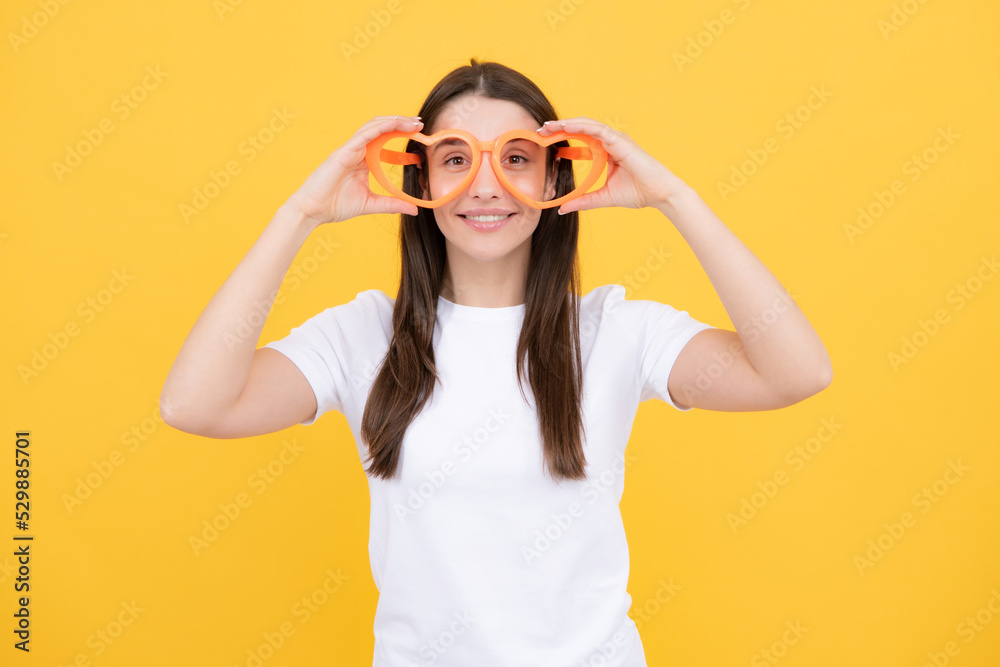 Portrait of a funny girl wearing cool party glasses. Cheerful young girl smiling with heart-shape glasses on yellow background.