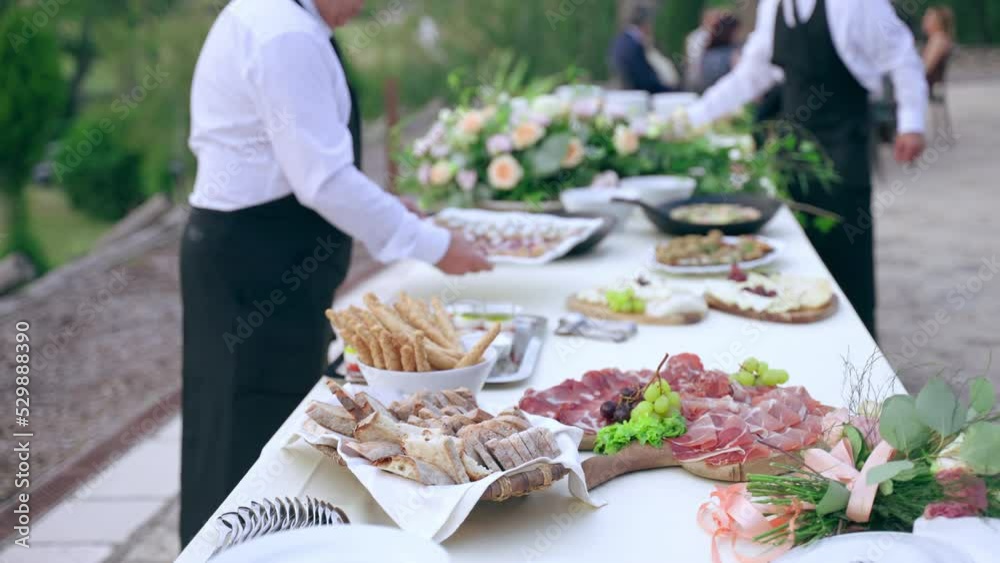 High-qualified restaurant waiters in uniform serving official exit party, beautifully decorated banquet table prepared for invited business guests. Exclusive catering food on silver trays, plates and