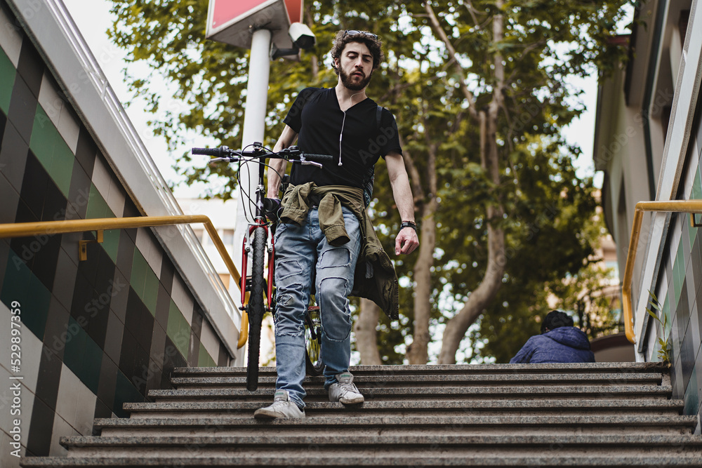 A young hipster guy descends the stairs to the subway station carrying a bicycle - lifestyle concept of people, city transport and urban sustainability A young hipster guy descends the stairs to the subway station carrying a bicycle - lifestyle concept of people, city transport and urban sustainability