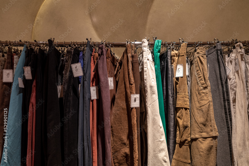 Row of earth tone colour fabric jeans or denim trousers and slacks hang on hanger. Various trousers in retail fashion store. Second hand retro style of dark tone slacks sold in outlet shop.