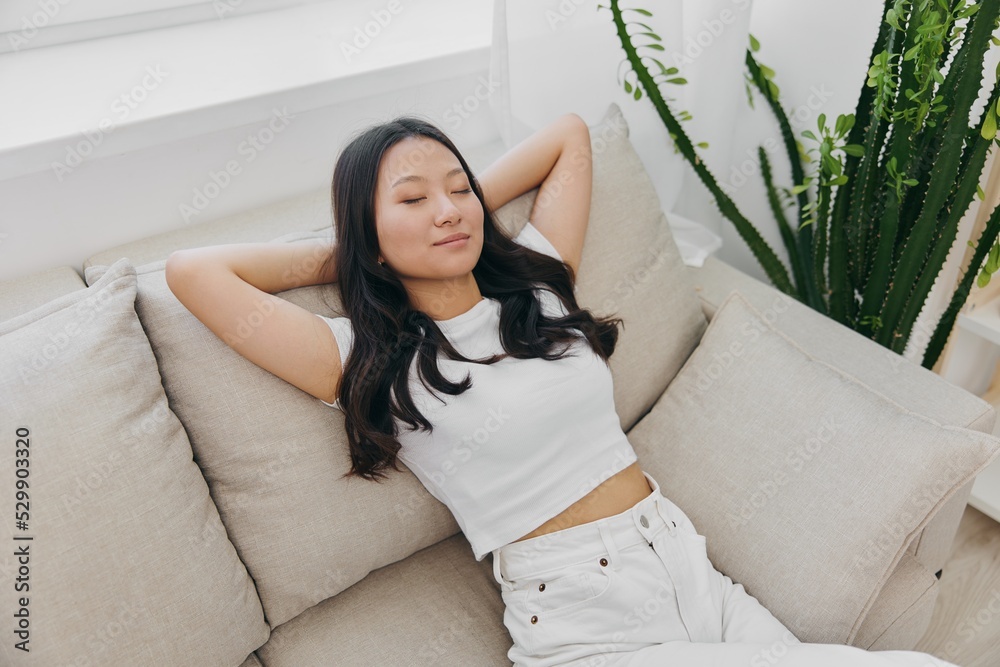 A young Asian woman sits at home on the couch resting asleep with her hands behind her head and smiling, waking up in the morning and drowsy during the day A young Asian woman sits at home on the couch resting asleep with her hands behind her head and smiling, waking up in the morning and drowsy during the day