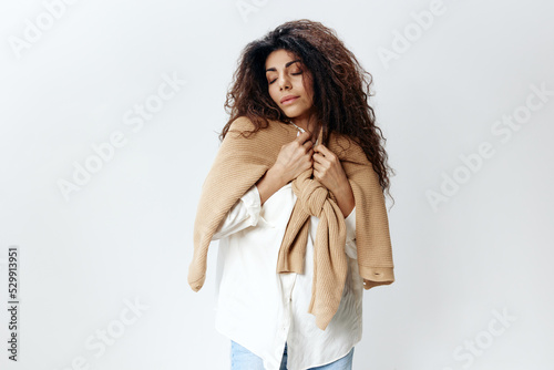 Curly Pretty Latin Female in White Shirt Enjoying Pastel Warm Sweater Cardigan Posing Isolated At White Studio Wall Background. Fashion Seasonal Sale New Collection Concept. Copy space