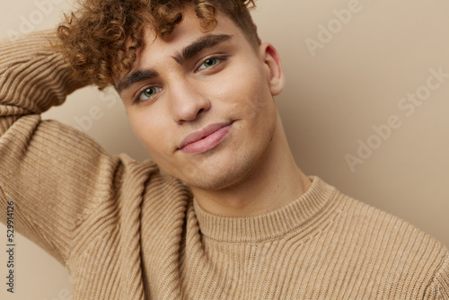 a handsome, attractive man with curly hair, in stylish clothes stands on a beige background and poses relaxed holding his hand by his hair, smiling pleasantly. Horizontal Studio Photography