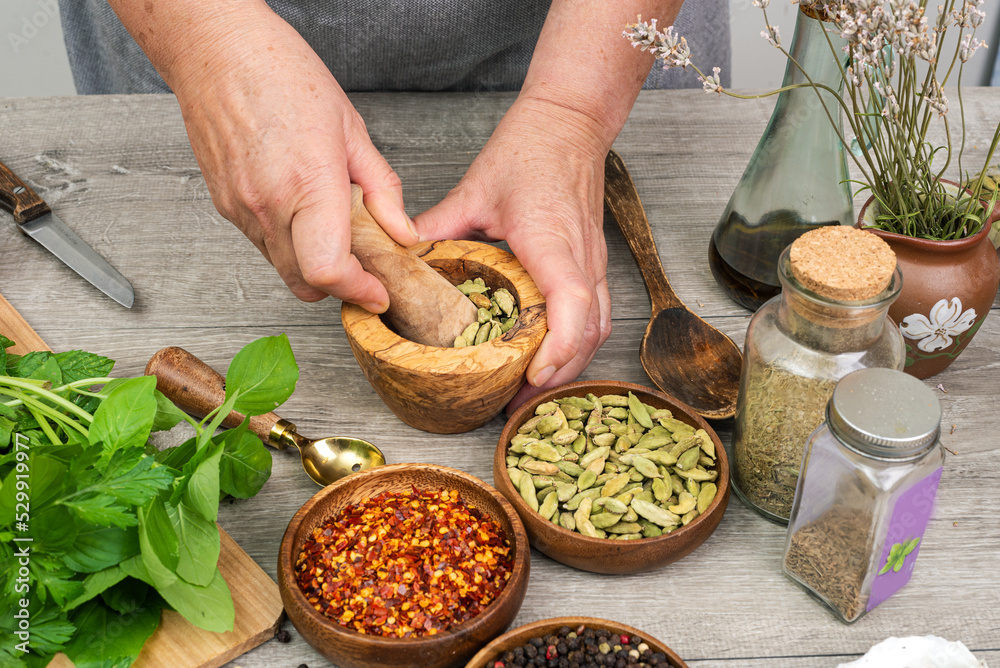 Woman's hands grind cardamom in a wooden mortar. Kitchen gray table with various spices. Woman's hands grind cardamom in a wooden mortar. Kitchen gray table with various spices.