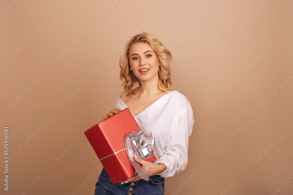 A smiling blonde woman in a white blouse holding a red gift box isolated on a beige background. Copy space. A smiling blonde woman in a white blouse holding a red gift box isolated on a beige background. Copy space.