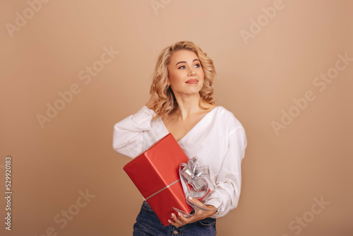 A smiling blonde woman in a white blouse holding a red gift box isolated on a beige background. Copy space. © VikaNorm