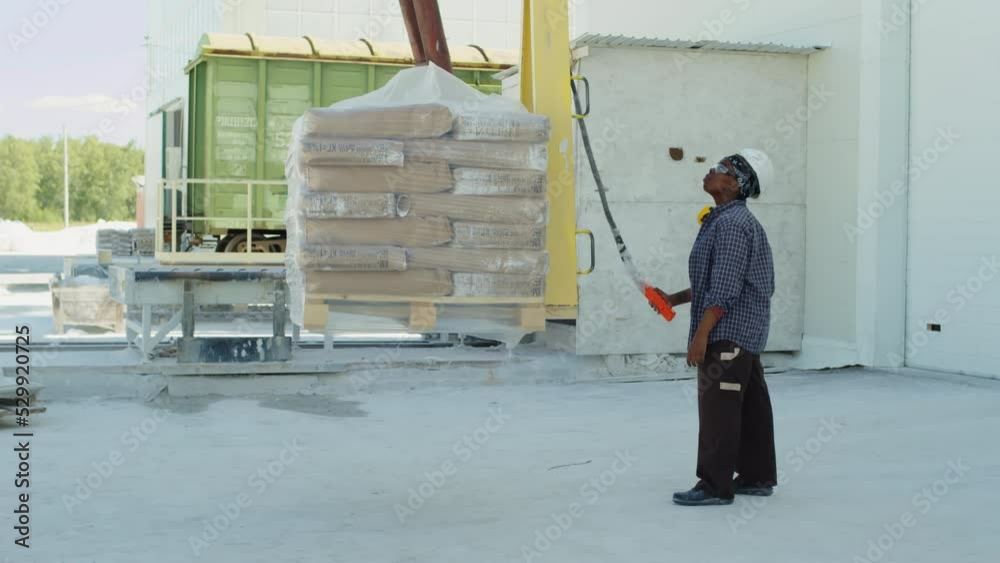 Full length shot of young female African American plant worker lifting heavy natural stone slabs on crane outside marble processing factory Full length shot of young female African American plant worker lifting heavy natural stone slabs on crane outside marble processing factory