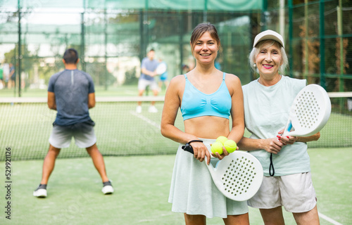 Portrait of friendly young and senior women standing on padel tennis court together during training outdoors. © JackF