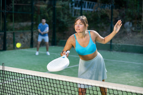 Portrait of emotional fit young woman playing padel tennis on open court in summer, swinging racket to return ball over net. Sportswoman ready to hit volley