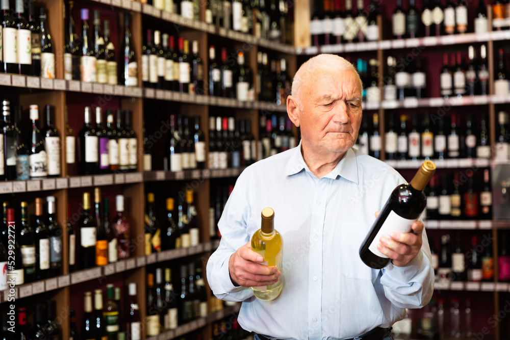 Serious elderly man chooses between red and white wine in a liquor store. High quality photo
