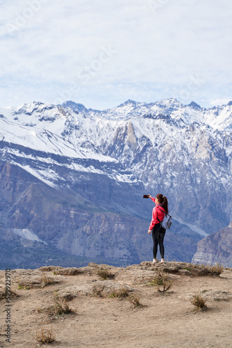 young red haired woman with red jacket and backpack  taking pictures with her phone in the middle of the Andes mountain range in Chile