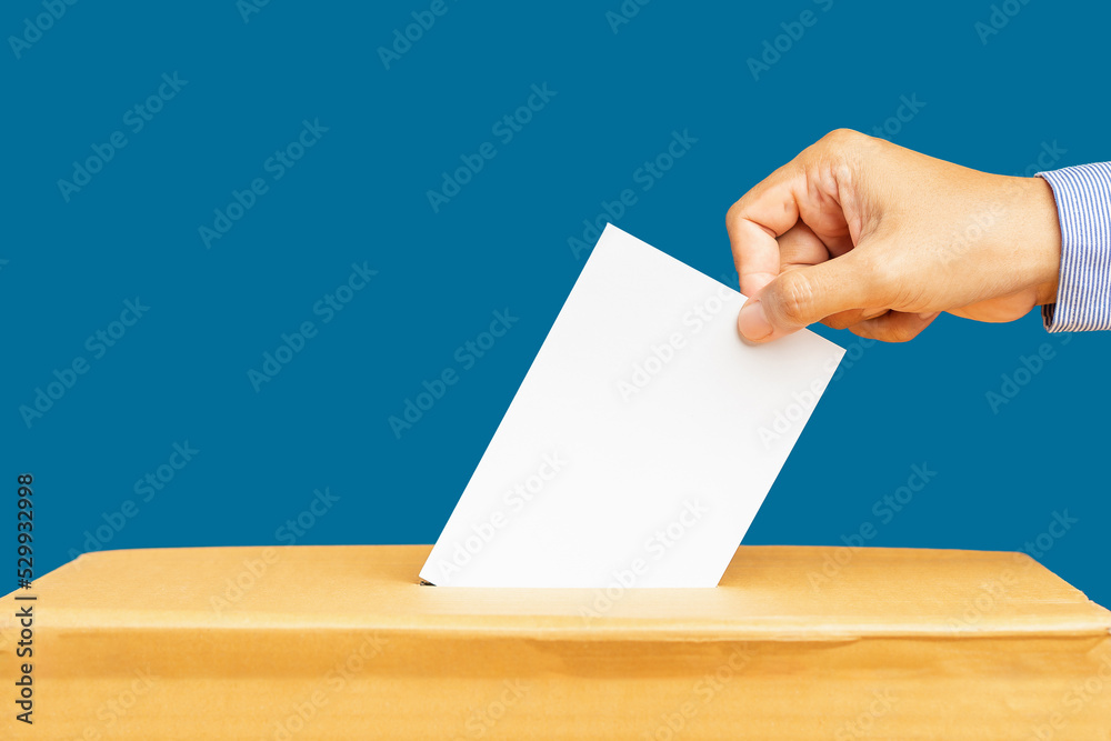 Hand voter holding ballot paper putting into the voting box at place election against a blue background Hand voter holding ballot paper putting into the voting box at place election against a blue background