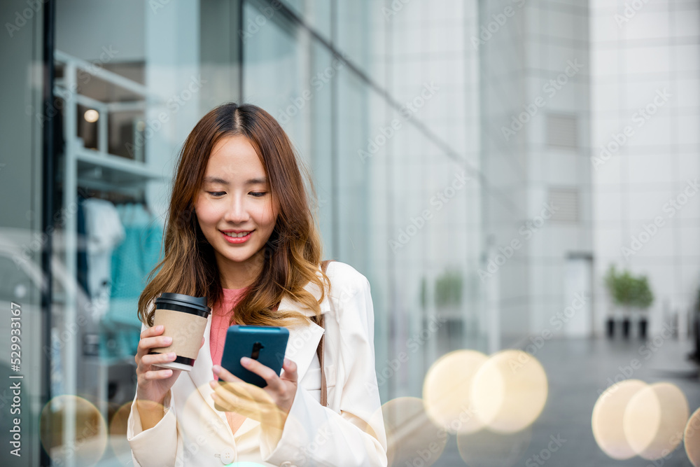 Asian businesswoman holding coffee cup takeaway look smartphone going to work walk on city street near office in morning, Portrait business woman hold mobile phone and paper cup of hot drink outdoor