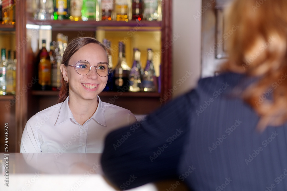 Cheerful bartender woman talking to client in cafe. Positive young female in casual clothes and eyeglasses smiling standing at counter in modern bar