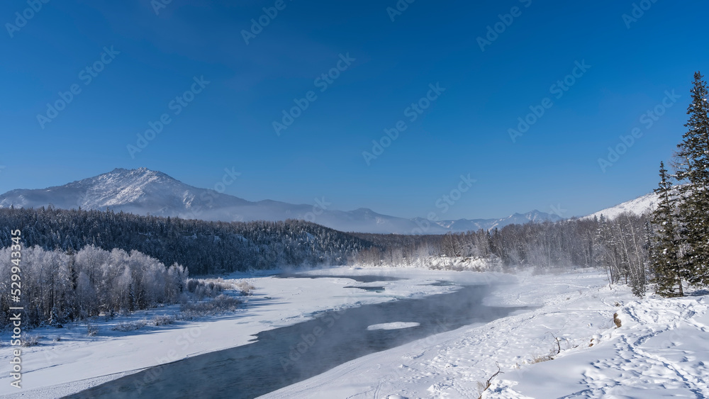 An ice-free river in a snow-covered valley. Steam rises above the water.  Footprints in snowdrifts. The trees in the forest are covered with hoarfrost. A mountain against a clear blue sky. Copy space