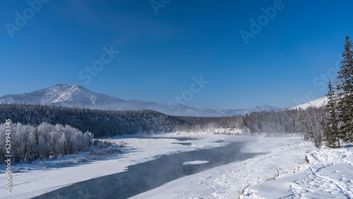 An ice-free river in a snow-covered valley. Steam rises above the water. Footprints in snowdrifts. The trees in the forest are covered with hoarfrost. A mountain against a clear blue sky. Copy space