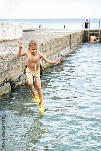Small boy jumps from stone pier to sea against people. Moment of excited child jumping and touching water. Small kid looks like magician