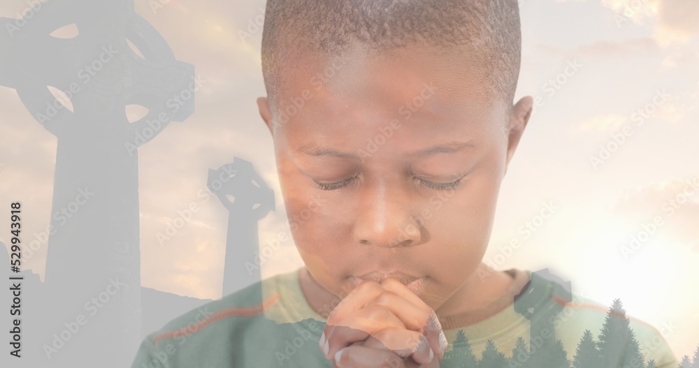Naklejka premium Multiple exposure of african american elementary boy praying with cemetery crosses against sky