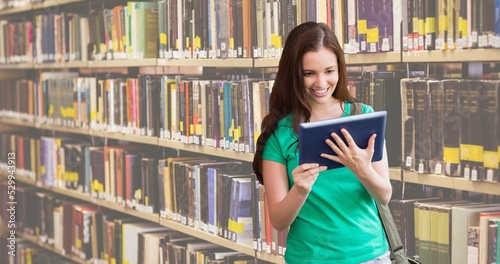 Smiling caucasian teenage girl reading book while standing against bookshelf in library