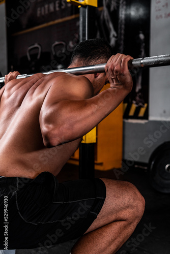 Man from behind lifting a barbell with weight on his shoulders. Shirtless Latino man lifting a heavy barbell. Flexed back of a muscular man. High quality photo