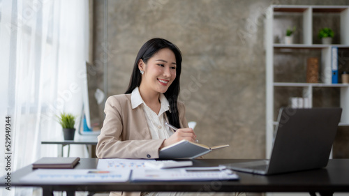 Beautiful Asian business woman using her laptop to work and enjoy working, taking note, reviewing assignment and smiling in the office.