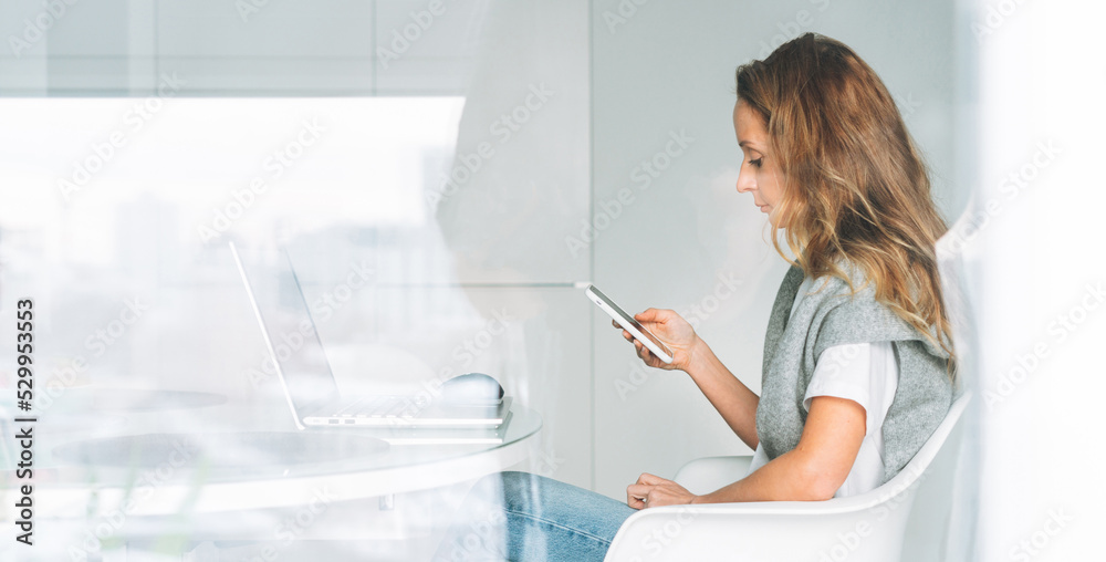 Young adult forty years beautiful blonde woman with long hair working on laptop using smartphone sitting on kitchen at home, remote work Young adult forty years beautiful blonde woman with long hair working on laptop using smartphone sitting on kitchen at home, remote work