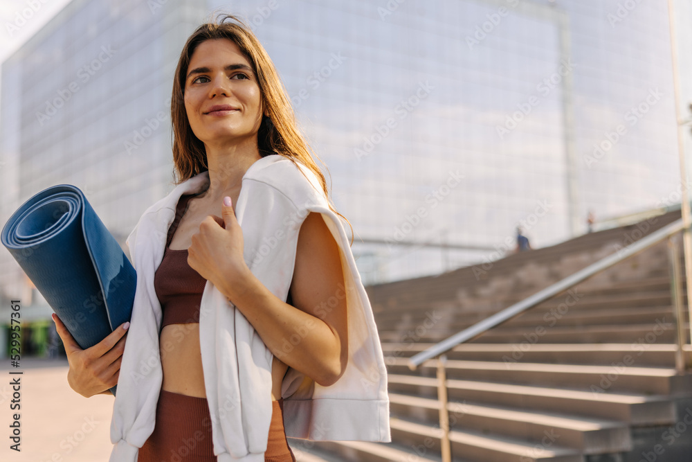 Fototapeta premium Charming beautiful young woman looking away, in hands blue mat. Staying on the stairs background at city and smiling. Concept of street style 
