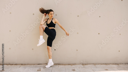 Full view of fit young woman keep leg bent while jumping on the wall background. Staying at street doing workout, looking away. Sport time concept 