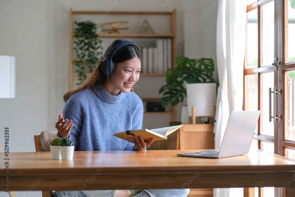 Smiling young asian teen girl wear headphones video calling on laptop. Happy pretty woman student looking at computer screen watching webinar or doing video chat by webcam. Smiling young asian teen girl wear headphones video calling on laptop. Happy pretty woman student looking at computer screen watching webinar or doing video chat by webcam.