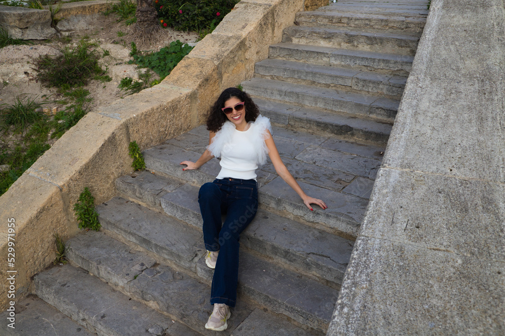beautiful young woman with dark, curly hair sitting on a staircase in a park. The woman wears sunglasses and makes different expressions. Expressions and lifestyle concept beautiful young woman with dark, curly hair sitting on a staircase in a park. The woman wears sunglasses and makes different expressions. Expressions and lifestyle concept