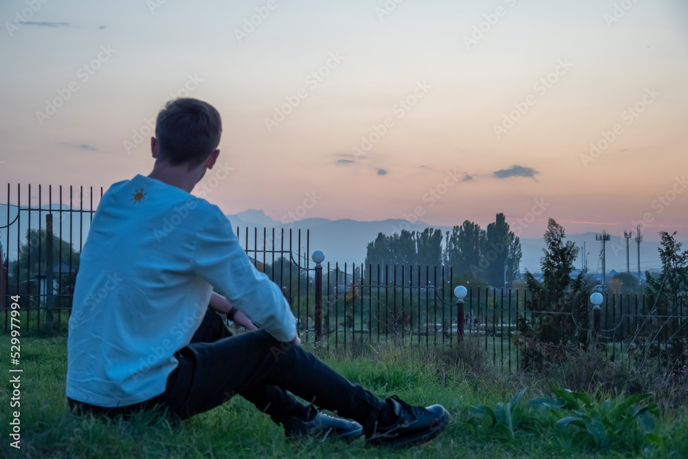 Fototapeta premium The guy sits on the grass admiring the sunset