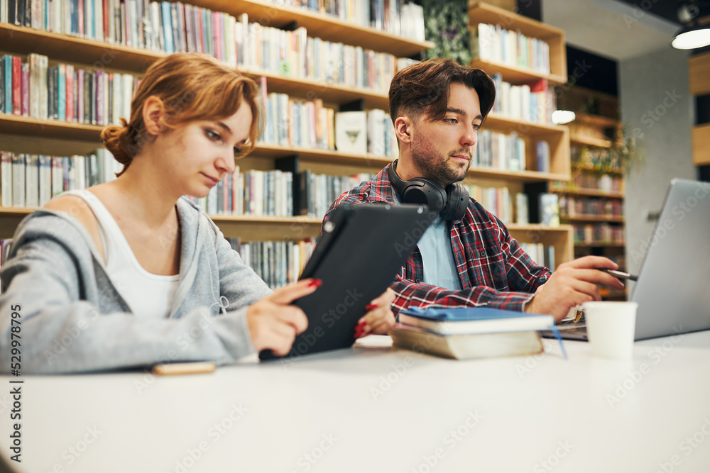 Students learning in university library. Young man preparing for test on laptop. Girl writing essay and making notes using computer. Focused students studying for college exams Students learning in university library. Young man preparing for test on laptop. Girl writing essay and making notes using computer. Focused students studying for college exams