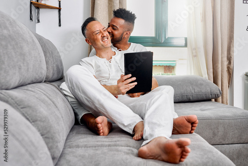 Young man biting ear of boyfriend sitting on sofa at home