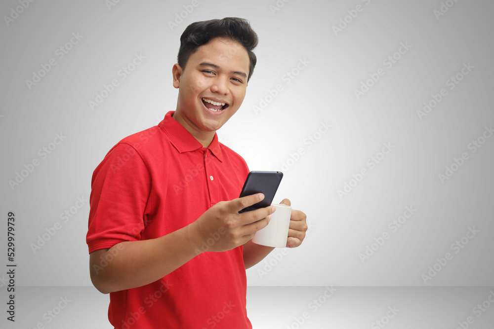 Portrait of cheerful Asian man in red polo shirt smiling at camera while holding mobile phone and a mug. Isolated image on gray background Portrait of cheerful Asian man in red polo shirt smiling at camera while holding mobile phone and a mug. Isolated image on gray background
