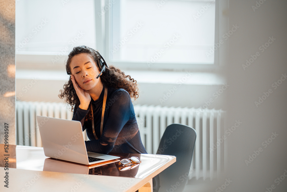 Young businesswoman wearing headset sitting with eyes closed at desk in office Young businesswoman wearing headset sitting with eyes closed at desk in office