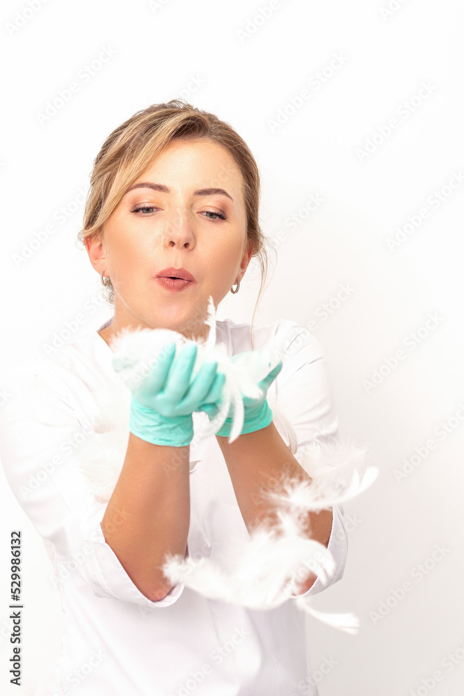 Young beautiful woman beautician in protective green gloves standing and blowing on feathers over white wall background