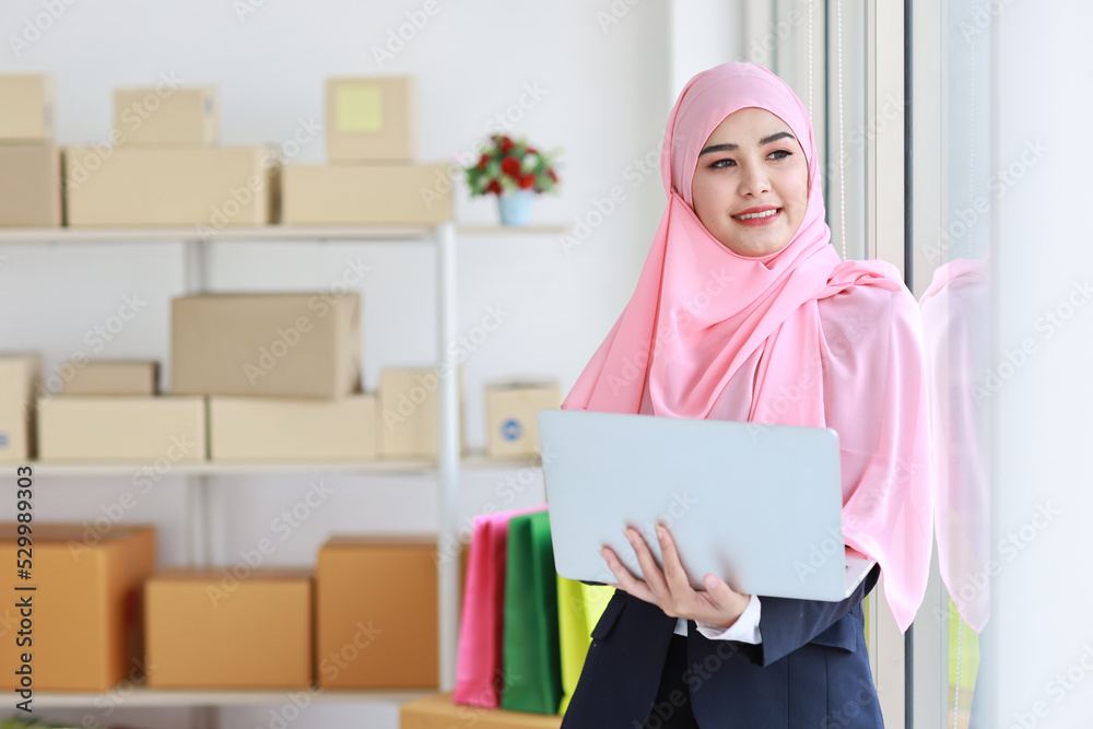 Religious asian muslim woman in blue suit and pink shaft on head standing and using computer with confidence. Business woman stand with package SME box delivery background. Work at home concept Religious asian muslim woman in blue suit and pink shaft on head standing and using computer with confidence. Business woman stand with package SME box delivery background. Work at home concept